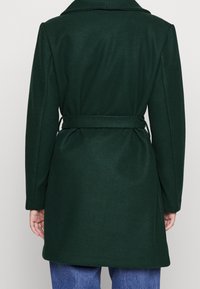 Mörkgrön yllekappa med bälte, långa ärmar, bred krage och strukturerad siluett. Slät textur utan synliga mönster.