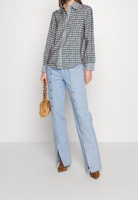 Camicia a maniche lunghe con motivo geometrico blu e marrone; pantaloni azzurri a vita alta con bottoni a pressione lungo i lati e spacchi.