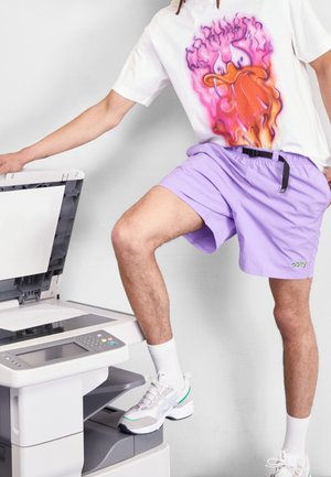 Purple shorts with a black belt and green logo; white t-shirt featuring a colorful graphic design; standing on a photocopier.