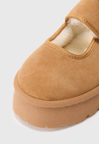 Lys brun semsket sko med rund tå, elastisk stropp og pelsfôr. Tyk, teksturert beige såle med sikksakk-mønster langs kantene.