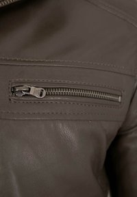Braune Lederjacke mit Details, die eine vertikale Reißverschlusstasche mit Metallreißverschluss, sichtbare Nähte und eine glatte, strukturierte Oberfläche aufweisen.