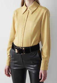 Gul langærmet bluse med krave, frontknapper og en brystlomme, parret med højtaljede sorte læderbukser og et sort bælte.