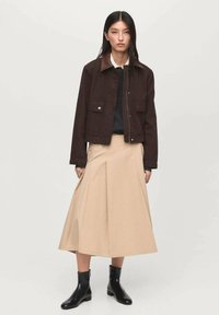 Veste marron avec un col contrastant, haut noir et jupe plissée beige, associée à des bottines noires. Tissu lisse et design structuré.