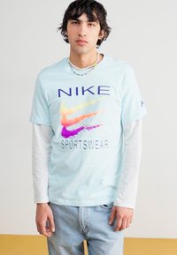 Šviesiai mėlynos spalvos trumpomis rankovėmis Nike marškinėliai su spalvingu logotipu ir tekstu "SPORTSWEAR", dėvimi ant baltų ilgomis rankovėmis marškinių. Dėvimos džinsinės kelnės.