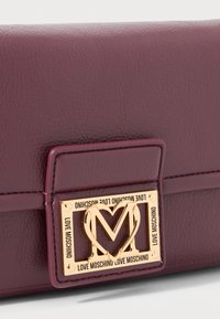 Close-up of purple leather purse flap with gold Love Moschino heart-shaped M logo clasp.