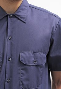 Korteblauwe shirt met korte mouwen in een gladde stof, voorzien van een puntkraag, twee knoopzakken op de borst en zwarte knopen. Eenvoudig ontwerp met minimale textuur.