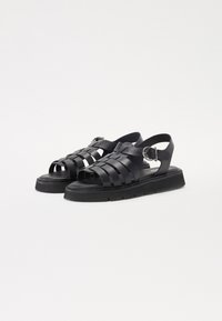 Sandale negre cu un design împletit, curea ajustabilă, talpă plată și material texturat. Prezintă o estetică îndrăzneață și contemporană.