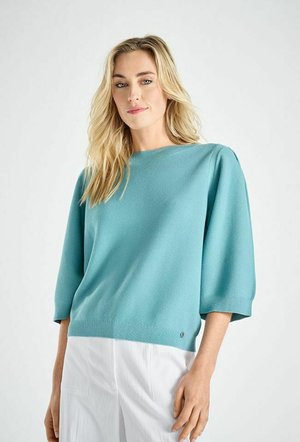 Blonde vrouw die een teal trui met driekwart mouwen en een witte broek draagt, staande tegen een effen lichte achtergrond.