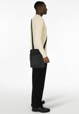 Schwarze Umhängetasche mit verstellbarem Riemen, glatter Textur, vorderer Reißverschlusstasche, Logodetail. Modell trägt cremefarbenes Oberteil und enge schwarze Hose.