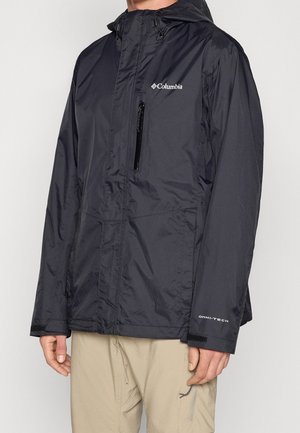 Man draagt een zwarte, waterdichte Columbia-jas met capuchon en beige broek, met zichtbare borstzak met rits en logo op de mouw.