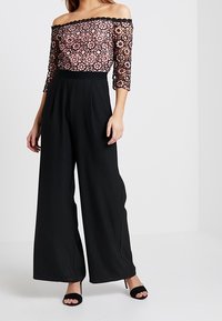Jumpsuit noir à larges jambes avec un haut en dentelle décolleté, arborant un motif floral rose. Texture lisse et ajustement réglable.