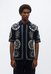 Πουκάμισο κοντομάνικο σε ναυτικό μπλε με κλασικό γιακά, διακοσμημένο με μοτίβα ήλιου σε εκρού, με κουμπιά μπροστά και υφή πλεκτού υφάσματος.