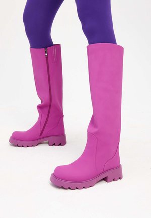 Bottes hautes violettes avec fermeture éclair latérale portées sur des collants violet vif, visibles de la moitié du mollet vers le bas sur fond blanc.