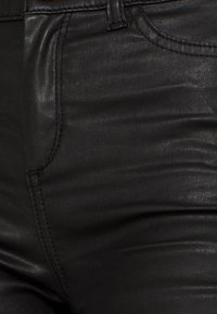 Gros plan sur un pantalon en cuir noir montrant la poche avant, la ceinture et le tissu texturé avec des détails de couture visibles.