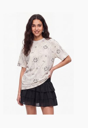 Ung kvinde med langt bølget hår iført en hvid T-shirt med sorte smilende ansigter og et sort lagdelt nederdel, smilende med den ene hånd på hoften.