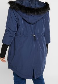 Granatowa kurtka puchowa z kapturem obszytym sztucznym futrem, ze ściągaczem w talii i długimi rękawami. Posiada zaokrąglony dół i boczne zamki. Gładka, wodoodporna tkanina.
