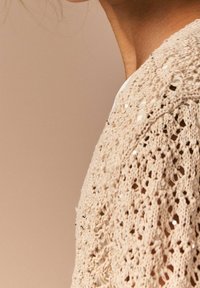 Strikket cardigan i lys beige med indviklet blondermønster og små pailletter, der viser tekstur og åben vævning på ærmerne.