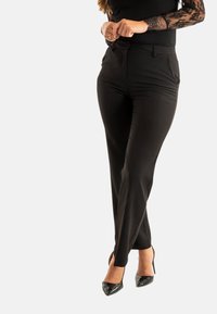 Zwarte tailored broek met een gladde textuur, met een standaard tailleband en details aan de zijnaad, gedragen met hoge zwarte schoenen.