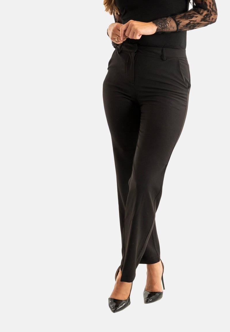 Zwarte tailored broek met een gladde textuur, met een standaard tailleband en details aan de zijnaad, gedragen met hoge zwarte schoenen.