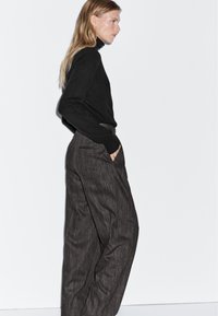 Zwarte coltrui gecombineerd met hoog aangesloten, donker gestreepte broeken. De outfit heeft een getailleerde silhoutte en zijzakken.