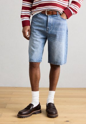 Mann trägt hellblaue Jeansshorts, braunen Ledergürtel, rot-weiß gestreiften Pullover, weiße Socken und dunkelbraune Loafers, steht auf einem Holzboden.