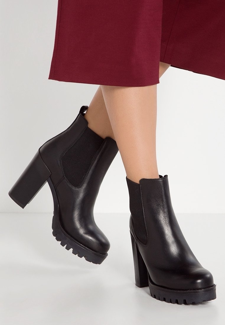 Femme portant des bottines en cuir noir à talons hauts avec des semelles épaisses et des panneaux latéraux élastiques, assorties à une jupe bordeaux.