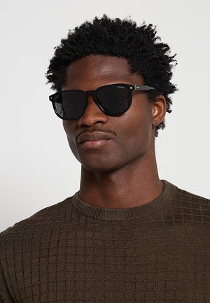 Uomo con capelli scuri e ricci che indossa occhiali da sole neri rettangolari e un maglione marrone lavorato a maglia su uno sfondo chiaro e uniforme.