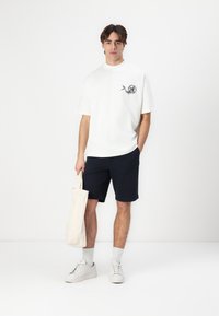 Hvit kortermet t-skjorte med grafisk trykk, kombinert med marineblå shorts. Modellen holder en kremfarget totebag og har på seg hvite joggesko og sokker.