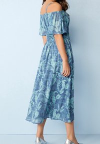 Vestido largo floral azul con mangas descubiertas, cintura fruncida y tejido ligero. Presenta un estampado de hojas verdes y unas bailarinas plateadas.