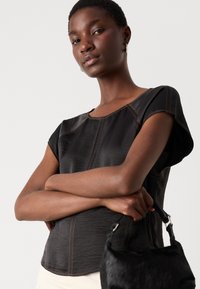 Schwarzes T-Shirt mit kurzen Ärmeln, strukturiertem Finish und orangefarbenen Nähten, kombiniert mit einer kleinen schwarzen Handtasche aus Fell.