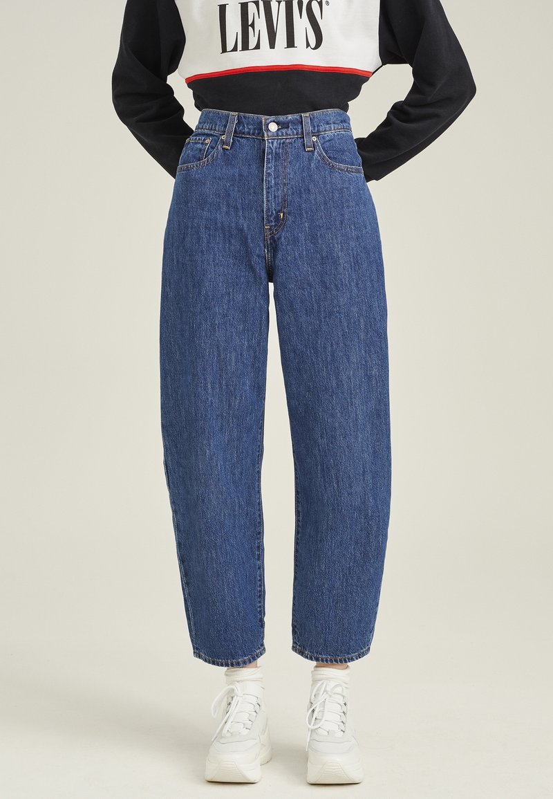 Personne portant un jean bleu taille haute, des baskets blanches et un sweat-shirt Levi's noir et blanc, debout les mains sur les hanches.