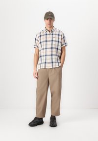 Camisa de manga curta xadrez em creme com linhas azuis e laranjas, combinada com calças largas bege e sapatos de desporto pretos. Apresenta um boné verde.