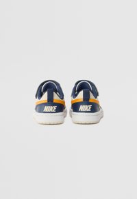 Tummansininen ja kreikkalainen Nike-lenkkari, jossa oranssit yksityiskohdat. Kenkä on pehmeä ja siinä on tarranauhat sekä teksturoitu valkoinen kumipohja.