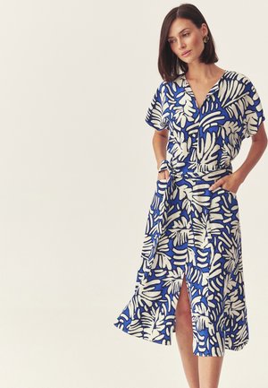 Vrouw met donker bobkapsel, gekleed in een blauw-witte jurk met een bladpatroon, zakken en een split aan de voorkant, staand met handen in de zakken.