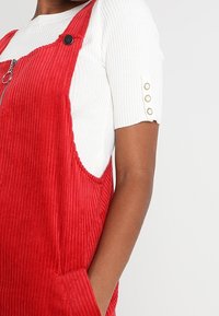 Rote Corduroy-Latzhose mit einem Reißverschluss vorne, seitlichen Taschen und schwarzen Knopf-Accents, kombiniert mit einem weißen gerippten T-Shirt mit kurzen Ärmeln.