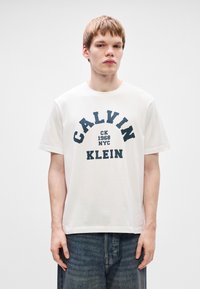 Valge puuvillane T-särk, millel on tumesinised graafilised tekstid "CALVIN KLEIN" ja "CK 1968 NYC". Särgil on lühikesed varrukad ja ümmargune kaelus.