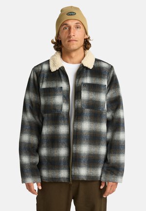 Man in a plaid jacket with a faux fur collar, wearing a beige beanie, stands against a white background, hands relaxed at his sides.