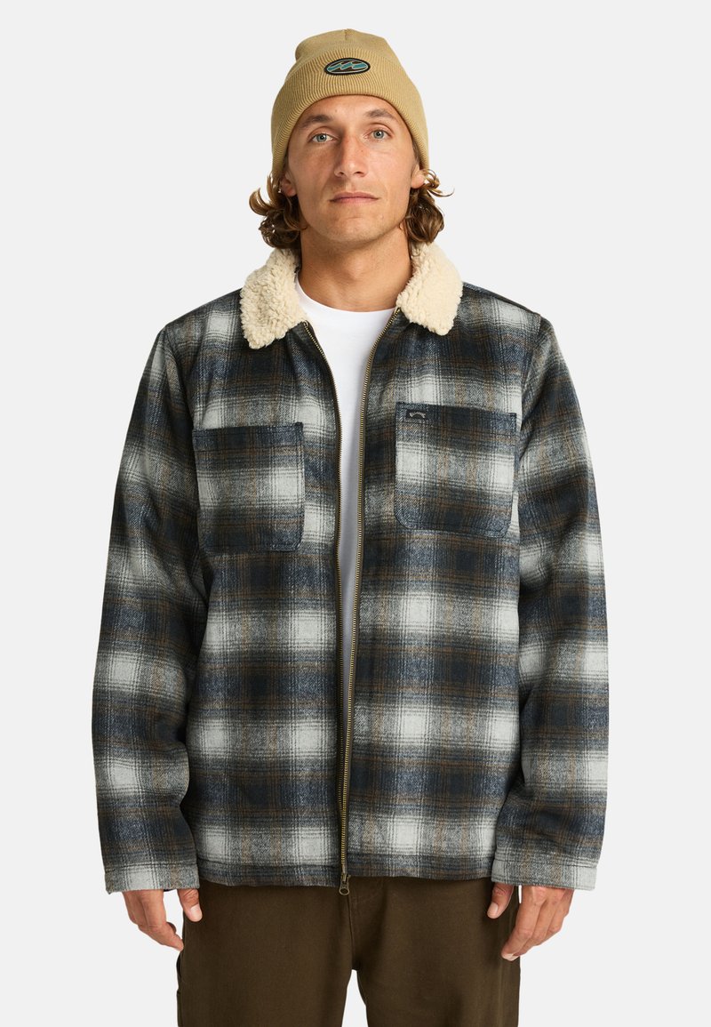 Man in een geruit jasje met een nepbont kraag, die een beige beanie draagt, staat tegen een witte achtergrond met zijn handen ontspannen langs zijn zij.