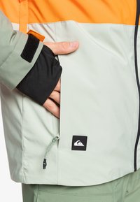 Chaqueta impermeable de color verde claro con acentos naranjas, puños ajustables, bolsillos con cremallera y un parche de logo negro cerca del dobladillo.