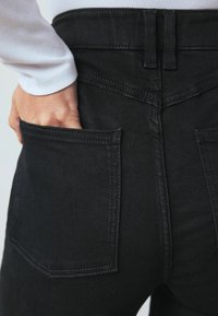 Fekete farmernadrág bemutatva, a hátsó zsebet kiemelve, varrás részletekkel és sima textúrával. Egy kéz a zsebben van.