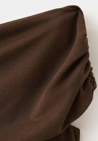 Tissu marron avec un détail froncé, probablement une manche ou une partie d’un vêtement, présenté sur un fond clair uni.