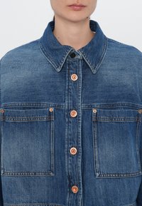 Jachetă din denim cu guler, nasturi din cupru, două buzunare pe piept și un finisaj albastru decolorat cu cusături vizibile.