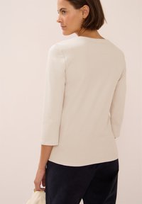 Ljus beige, figurnära topp med trekvartslånga ärmar och rund halsringning, parat med mörkblå byxor. Mjukt tyg, slät textur.