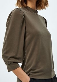 Mörk olivgrön topp med hög hals och puffade ärmar som når tre kvartar. Tillverkad av mjukt tyg med en slät yta.