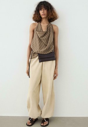 Femme aux cheveux bouclés portant un haut sans manches marron drapé, un pantalon beige à jambes larges, des sandales noires et une pochette noire à la taille devant un mur uni.