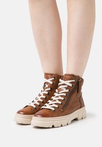 Bottes montantes en cuir marron avec des lacets blancs, fermeture éclair sur le côté, semelle en caoutchouc, finition texturée et semelle épaisse.
