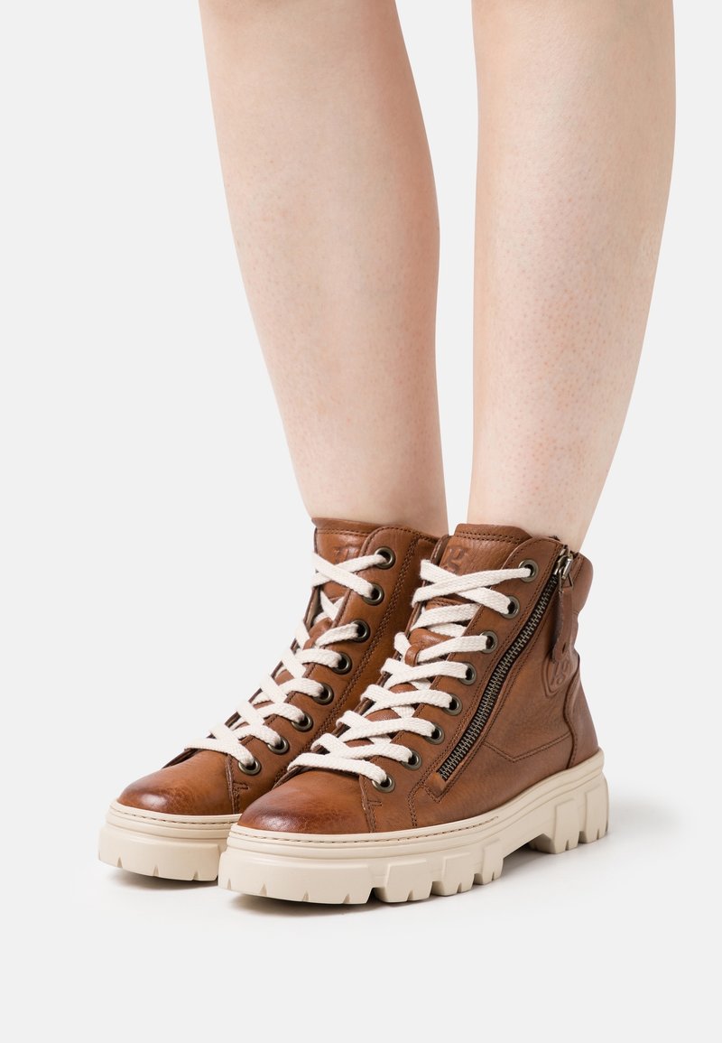 Bottes montantes en cuir marron avec des lacets blancs, fermeture éclair sur le côté, semelle en caoutchouc, finition texturée et semelle épaisse.
