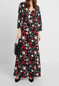 Maxikjol i svart tyg med ett blommönster i röd och vit. Har en V-ringning, långa ärmar och en knyts i midjan, kombinerad med en svart väska.
