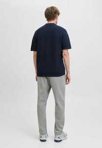 Uomo in piedi di spalle, indossa una maglietta blu scuro a maniche corte, pantaloni grigio chiaro e sneaker bianche su uno sfondo chiaro uniforme.