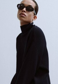 Czarny sweter dziany z wysokim kołnierzem i guzikami na jednym ramieniu, o prążkowanej fakturze i nowoczesnym kroju.